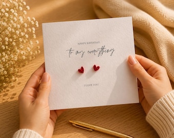 May include: A square white greeting card featuring the text HAPPY BIRTHDAY To my everything and I LOVE YOU in black ink. Two small red heart-shaped embellishments are attached to the center of the card between the printed text lines.