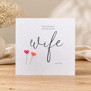 May include: A white greeting card with the word "wife" in cursive script, accompanied by the phrases "Happy Birthday to my beautiful" and "I love you." Two small, heart-shaped embellishments in pink and orange are below the word "wife."