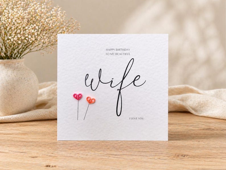 May include: A white greeting card with the word "wife" in cursive script, accompanied by the phrases "Happy Birthday to my beautiful" and "I love you." Two small, heart-shaped embellishments in pink and orange are below the word "wife."