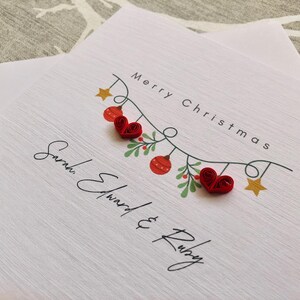 May include: White Christmas card with the text "Merry Christmas" and the names "Sarah Edward & Ruby". The card features a garland with red heart ornaments, red and green berries, and gold stars.