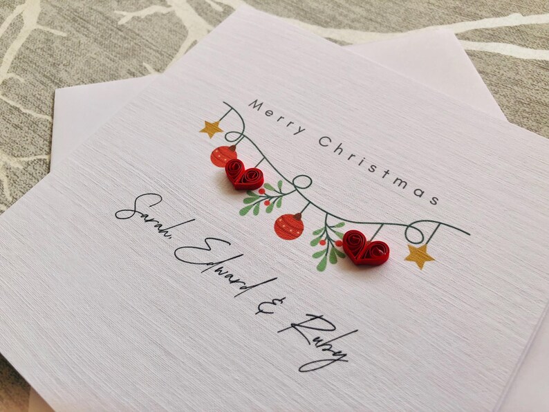 May include: White Christmas card with the text "Merry Christmas" and the names "Sarah Edward & Ruby". The card features a garland with red heart ornaments, red and green berries, and gold stars.