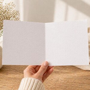 May include: An open, blank greeting card held by a person, with a textured white surface. A vase with dried flowers sits to the left. The card is on a wooden surface, with a soft, beige fabric in the background.