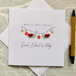 May include: A white Christmas card with the words "Merry Christmas" and a decorative garland of red ornaments, hearts, and stars. The names "Sarah Edward & Ruby" are written below the garland. A pen rests on the right side of the card.
