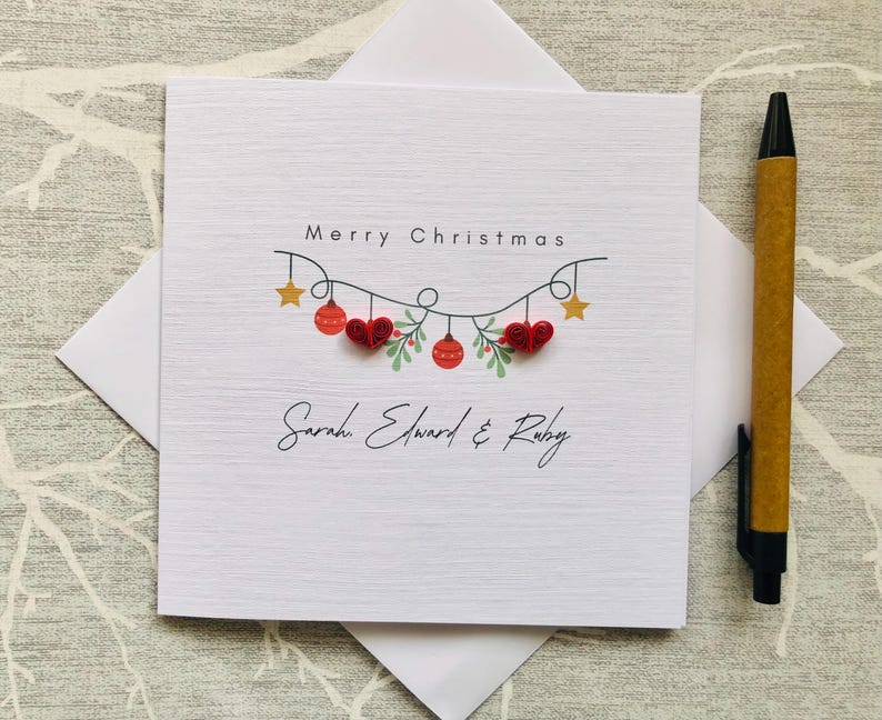 May include: A white Christmas card with the words "Merry Christmas" and a decorative garland of red ornaments, hearts, and stars. The names "Sarah Edward & Ruby" are written below the garland. A pen rests on the right side of the card.
