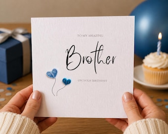 May include: A square white birthday greeting card with the text TO MY AMAZING Brother ON YOUR BIRTHDAY in black lettering. The card features two small blue paper quilled hearts with thin black stems attached to the lower left side of the text.