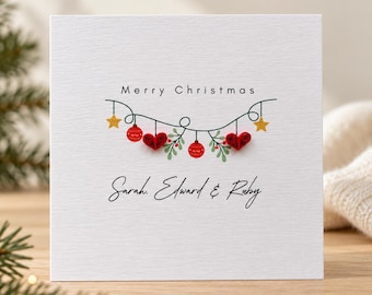 May include: A square white greeting card featuring the text Merry Christmas and the names Sarah, Edward & Ruby. The design includes a string of lights with yellow stars, red paper quilled ornaments, red paper quilled hearts, and green leaves.