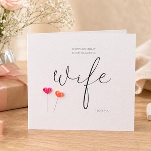 May include: A white greeting card with the word "wife" in elegant script. The card features the text "HAPPY BIRTHDAY TO MY BEAUTIFUL" and "I LOVE YOU". Two heart-shaped decorations are below the word "wife". A gift box and flowers are in the background.