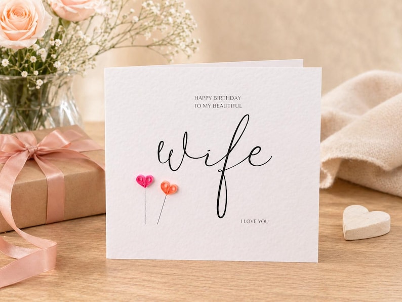 May include: A white greeting card with the word "wife" in elegant script. The card features the text "HAPPY BIRTHDAY TO MY BEAUTIFUL" and "I LOVE YOU". Two heart-shaped decorations are below the word "wife". A gift box and flowers are in the background.