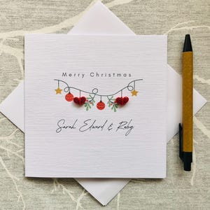 May include: A white Christmas card with the words "Merry Christmas" and a garland of red ornaments, hearts, and stars. The names "Sarah, Edward & Ruby" are written below. A pen rests on the side.