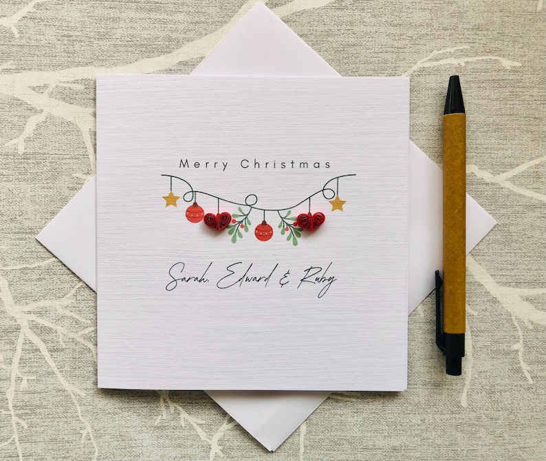 May include: A white Christmas card with the words "Merry Christmas" and a garland of red ornaments, hearts, and stars. The names "Sarah, Edward & Ruby" are written below. A pen rests on the side.