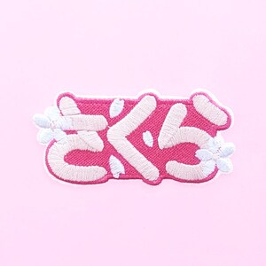 Könnte beinhalten: Ein pinkfarbener, gestickter Aufnäher mit japanischen Schriftzeichen und weißen floralen Akzenten. Der Aufnäher zeigt das japanische Wort "Sakura" in Weiß, umrandet in Pink, vor einem hellrosa Hintergrund. Der Aufnäher ist wahrscheinlich für Kleidung oder Accessoires.