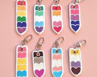 May include: Set of 8 keychains with rainbow heart designs. Each keychain features a different color combination of rainbow hearts. The keychains are made of fabric and have silver metal rings.