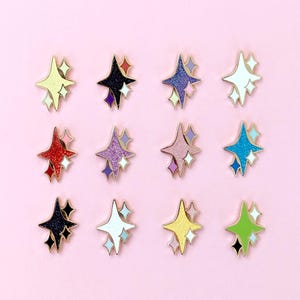May include: A collection of twelve star-shaped enamel pins in various colors, including yellow, black, purple, red, pink, and blue. Each pin features a gold outline and a smaller star accent. The pins are arranged on a light pink background.