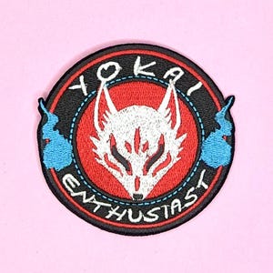 May include: A black and red embroidered patch with a white fox head in the center. The fox has red eyes and a white muzzle. The text "Yokai Enthusiast" is written in a circle around the fox head.