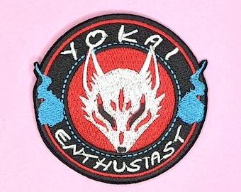 Peut inclure: Un écusson brodé noir et rouge avec une tête de renard blanche au centre. Le renard a les yeux rouges et un museau blanc. Le texte "Yokai Enthusiast" est écrit en cercle autour de la tête du renard.