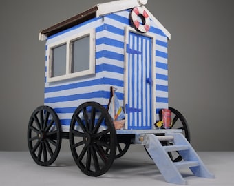Pode incluir: Uma cabana de praia azul e branca listrada sobre rodas com um salva-vidas vermelho no telhado. A cabana tem uma porta azul e uma pequena janela. Há um pequeno degrau de madeira que leva à porta.