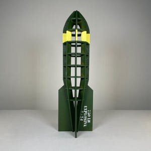 May include: A green wooden bomb-shaped sculpture with yellow detailing. The sculpture has a hollow, open design with horizontal supports. White text on the side reads "750 LB EXPLOSIVE 7-74".