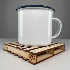 May include: A white enamel mug with a blue rim sits on a small wooden pallet coaster with the word "EPAL" printed on it.