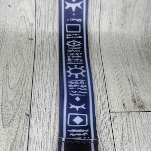 Op de afbeelding: Een donkerblauwe lanyard met witte geometrische patronen en symbolen. De lanyard heeft een zilverkleurige metalen sluiting en een splitring. Het ontwerp herhaalt zich over de gehele lengte.