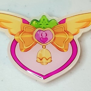 May include: A pink heart-shaped charm with a gold bow, green leaves, and a gold bell. The charm has wings on either side.