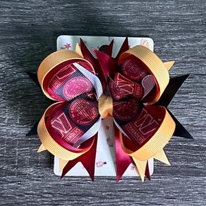 May include: A handmade hair bow with a layered design in burgundy, gold, and black ribbons. The bow features a printed ribbon with the text "COMMANDERS" and a logo. The bow is attached to a decorative card.