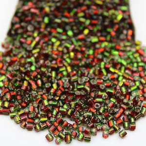 May include: A pile of red and green seed beads with a metallic sheen. The beads are small and cylindrical.