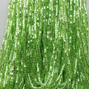 May include: Close-up of a collection of bright green, rectangular beads. The beads are strung together, creating a textured, flowing appearance. The beads have a reflective, metallic sheen.