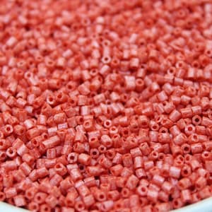 May include: Close-up of a bowl filled with small, cylindrical, coral-colored beads. The beads are uniform in size and shape, with a hole running through the center of each. The beads are densely packed, creating a textured surface. Ideal for jewelry making and crafts.