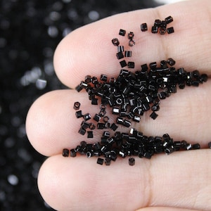 May include: Close-up of small, black, square-shaped beads. The beads are scattered across a person's open palm, with a larger quantity of the beads visible in the background. The beads appear to be made of a shiny material.
