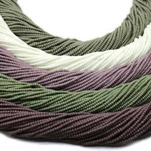 Puede incluir: Primer plano de una colección de cuentas de semillas en tres colores: blanco, verde claro y malva. Las cuentas están dispuestas en un patrón circular, creando un efecto de capas.