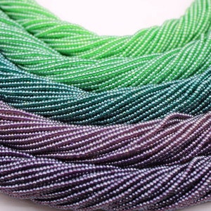 Puede incluir: Primer plano de un montón de cuentas de semillas iridiscentes en tonos verdes y morados. Las cuentas están dispuestas en un patrón circular, creando una pantalla texturizada y colorida.