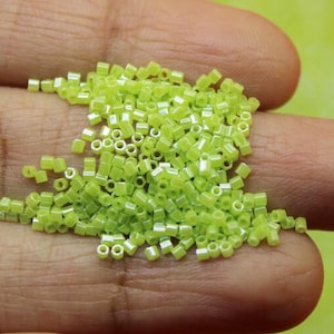 May include: A handful of small, light green, square-shaped beads. The beads are scattered across the fingers and palm of a hand, with a soft, blurred green background. These beads are likely used for jewelry making or other craft projects.
