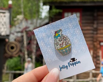 May include: A blue and white cupcake pin with white frosting and a snowflake design. The pin is on a card with a snowflake pattern and the text "Jolly Poppin".