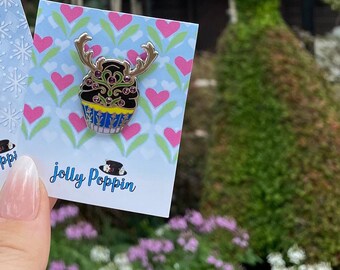 May include: A black and gold cupcake pin with a reindeer design and red and green accents. The pin is on a card with pink hearts and the text "Jolly Poppin".