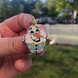 May include: A silver pin shaped like a snowman cupcake. The snowman has a carrot nose, black eyes, and a yellow smile. The cupcake is decorated with blue frosting and white snowflakes.