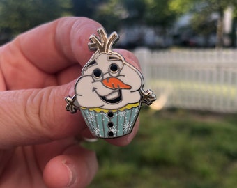May include: A silver pin shaped like a snowman cupcake. The snowman has a carrot nose, black eyes, and a yellow smile. The cupcake is decorated with blue frosting and white snowflakes.