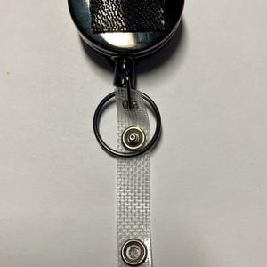 May include: A retractable badge reel with a shiny, silver-colored casing. A black mesh square is adhered to the top. A clear, woven strap with a metal snap and a metal ring are attached to the reel.