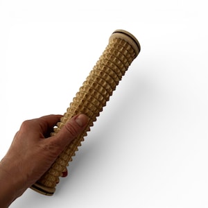 May include: A cylindrical wooden massage roller featuring a series of raised, textured ridges along its length. The tool has a light tan natural wood grain finish with flat circular end caps and a dark brown accent line on each end.