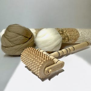 Peut inclure: Un outil de feutrage en bois avec une poignée et une roue à pointes. L'outil est utilisé pour feutrer les fibres de laine ensemble. Il y a deux boules de laine en arrière-plan, une brune et une blanche.