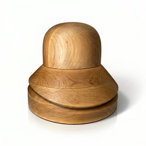 May include: A light brown wooden hat block featuring a smooth, rounded dome top section resting on a wider, tiered base with visible natural wood grain patterns throughout the structure, creating a solid, multi-layered cylindrical form.