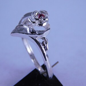 May include: A silver ring with a rose design and a small red gemstone in the center.