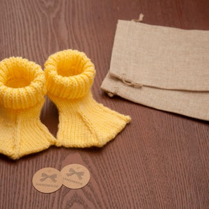 May include: Two yellow knitted baby booties with handmade tags. The tags are brown with white text and a white ribbon.