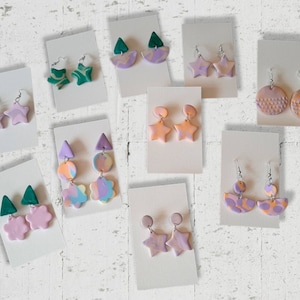 May include: Assortment of handmade polymer clay earrings in various shapes and colors, including stars, flowers, triangles, and circles. The earrings are displayed on white cards with silver hooks.