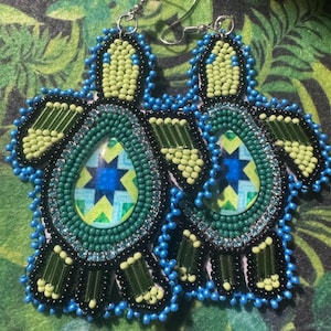 May include: A pair of beaded turtle earrings. The earrings feature a turtle shape with a central oval design in shades of green, blue, and yellow. The edges are outlined with black and blue beads, and the earrings have silver hooks.