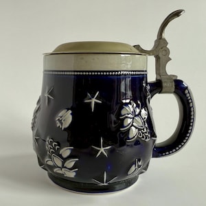 May include: A dark blue ceramic beer stein with white floral and star designs. It has a metal hinged lid and handle. The stein is approximately 7 inches tall.