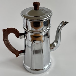 Può includere: Una caffettiera cromata vintage con manico e pomolo in legno marrone scuro. La caffettiera ha un beccuccio curvo e un manico a forma di cuore. È un classico articolo per la cucina.