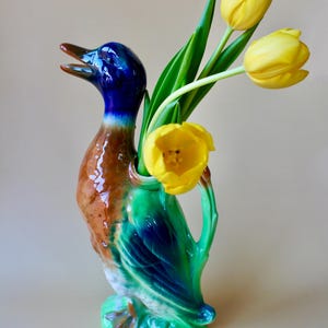 May include: A ceramic duck vase with a blue head, brown body, and green wings. The vase holds three yellow tulips with green stems and leaves. The vase is a decorative item.