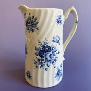 May include: A white ceramic pitcher with a blue floral pattern. The pitcher has a twisted design and a handle.