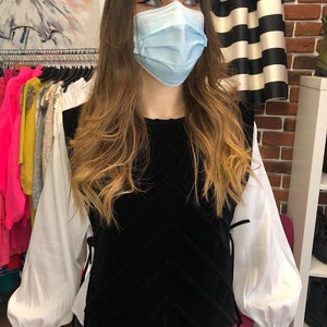 Op de afbeelding: Een zwart fluwelen vest met een chevron-patroon en een wit blouse met lange mouwen en gepofte schouders. Het vest heeft een strik aan de zijkant.
