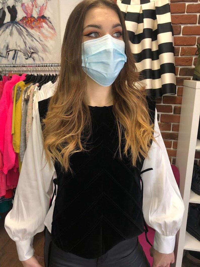 Op de afbeelding: Een zwart fluwelen vest met een chevron-patroon en een wit blouse met lange mouwen en gepofte schouders. Het vest heeft een strik aan de zijkant.